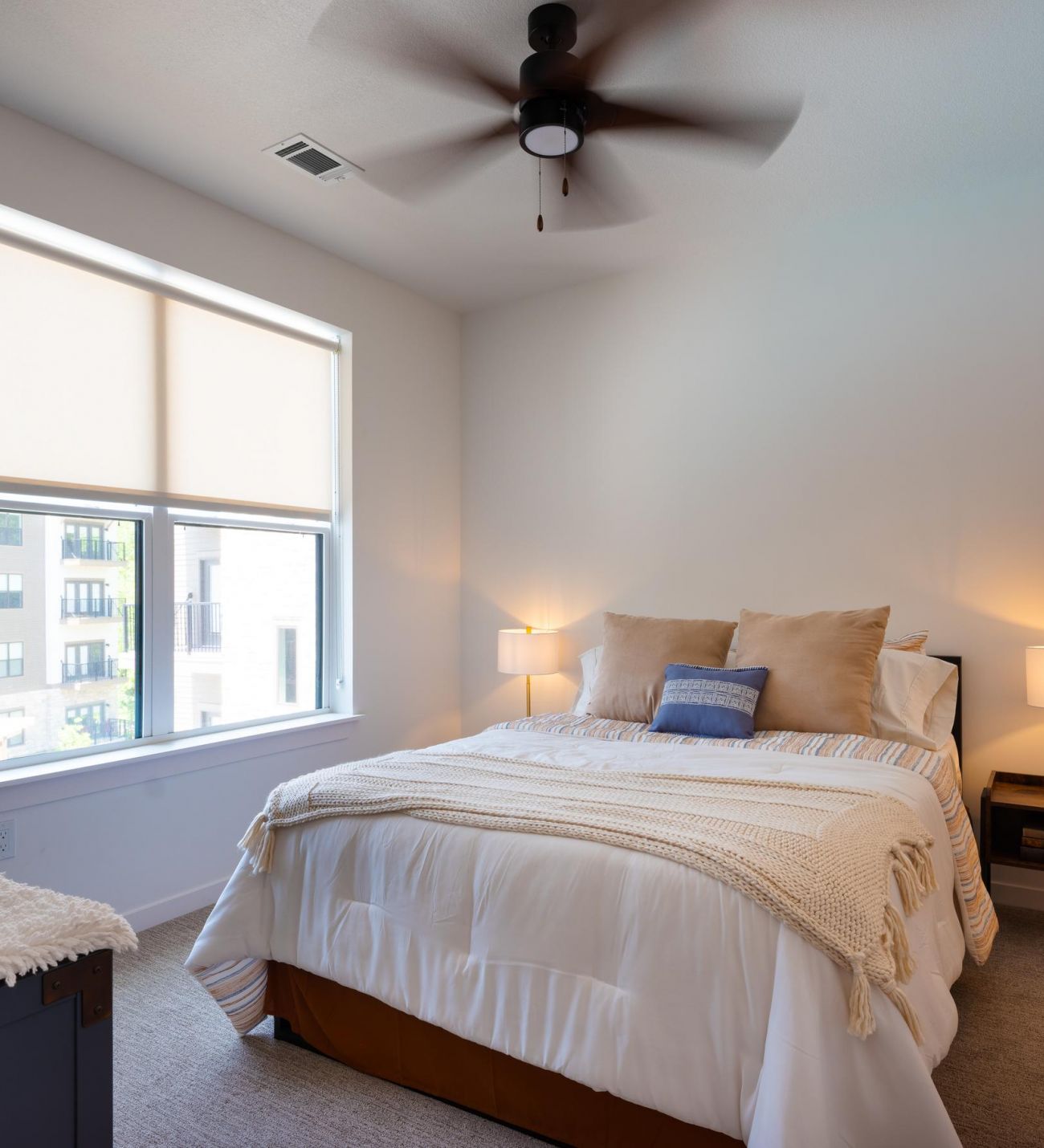 A cozy bedroom at Wheatfield Village featuring a queen-sized bed, large window with blinds, and a ceiling fan.
